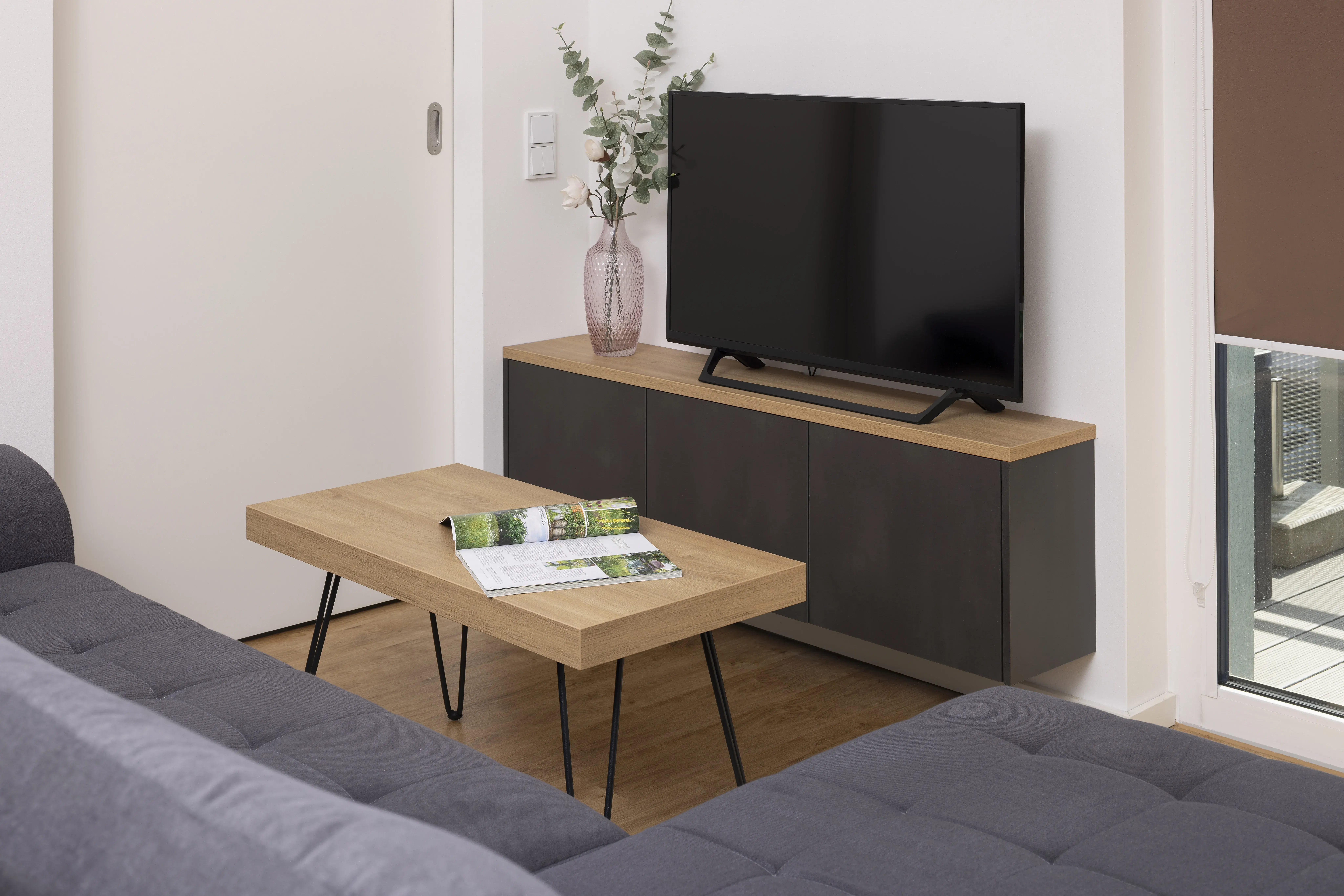 Modernes Wohnzimmer mit grauem Sofa, Holztisch, Fernseher und Vase mit Blumen.